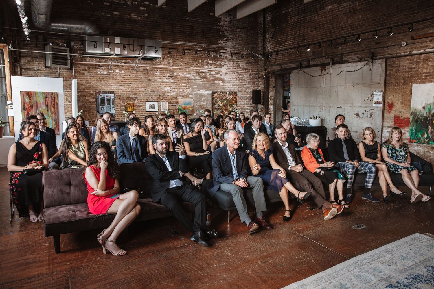 Wedding guests seated in a rustic brick-walled art gallery smile during a same-sex wedding ceremony.