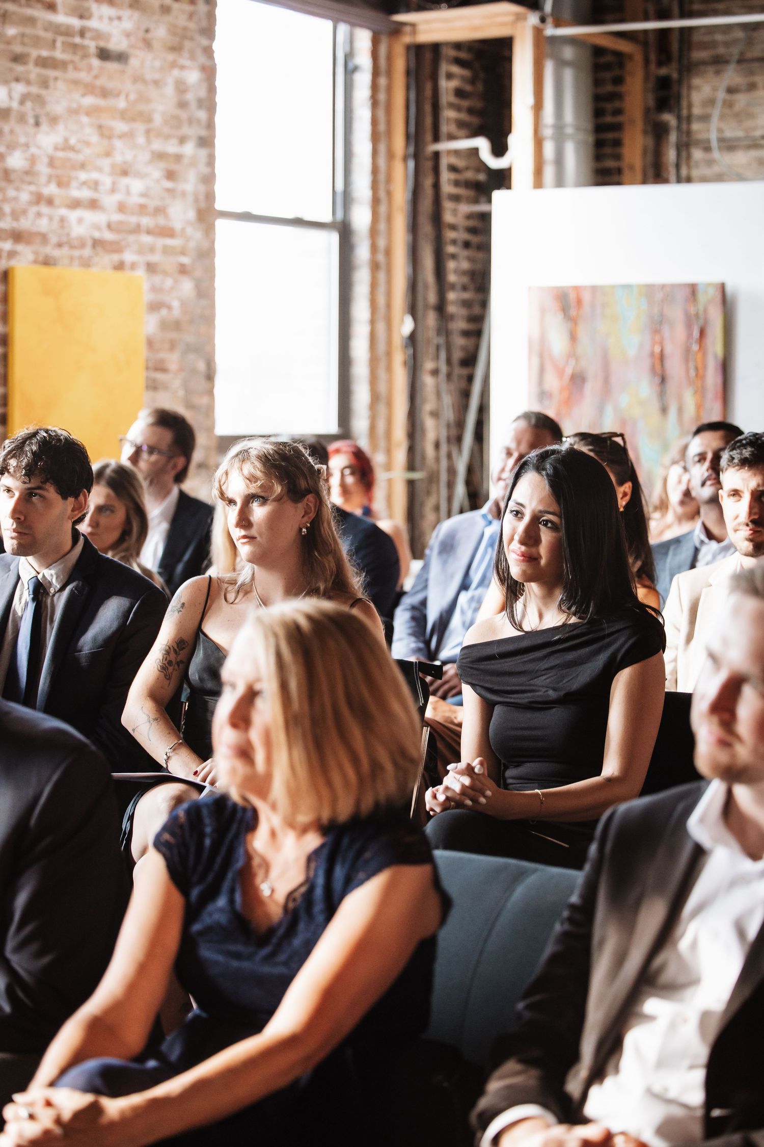 Diverse audience seated in rows at a formal art gallery event with brick walls.