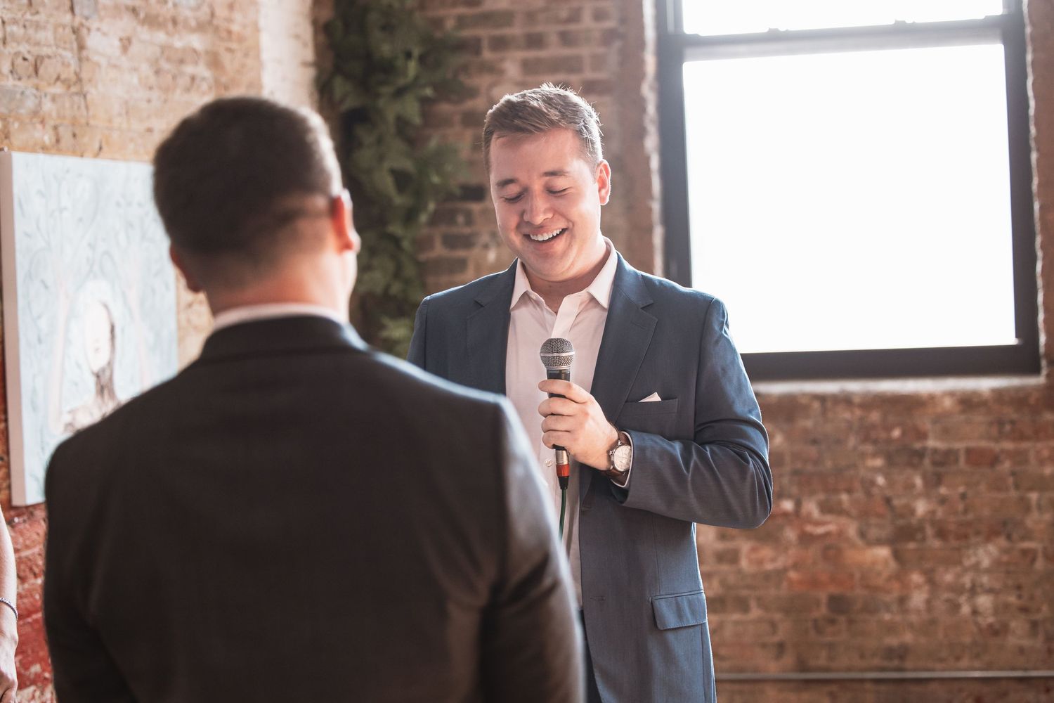 Smiling man in blue suit speaks into microphone at a sunlit brick-walled venue.
