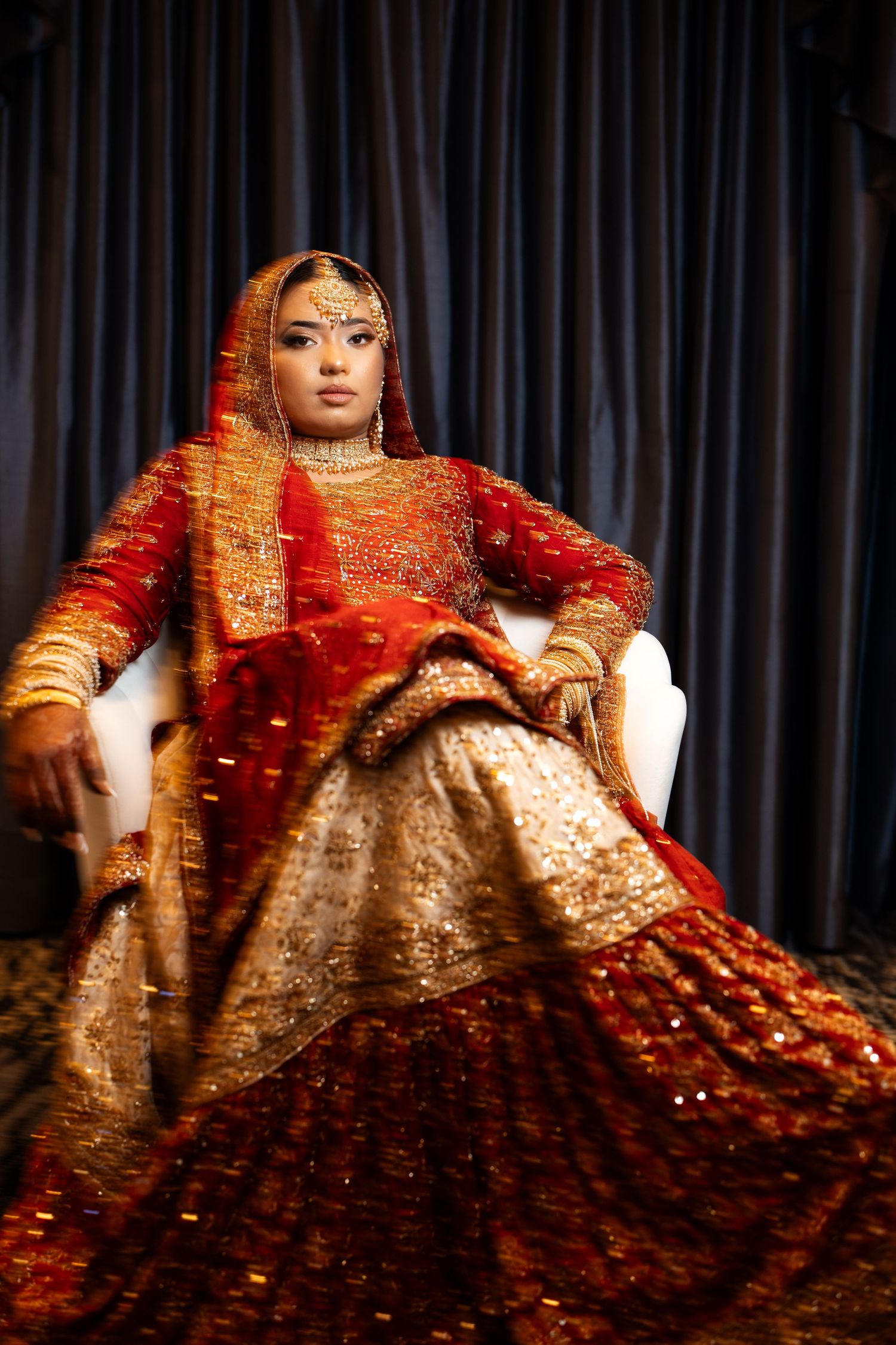 Bride in red and gold traditional lehenga sits gracefully in white chair against dark drapes.