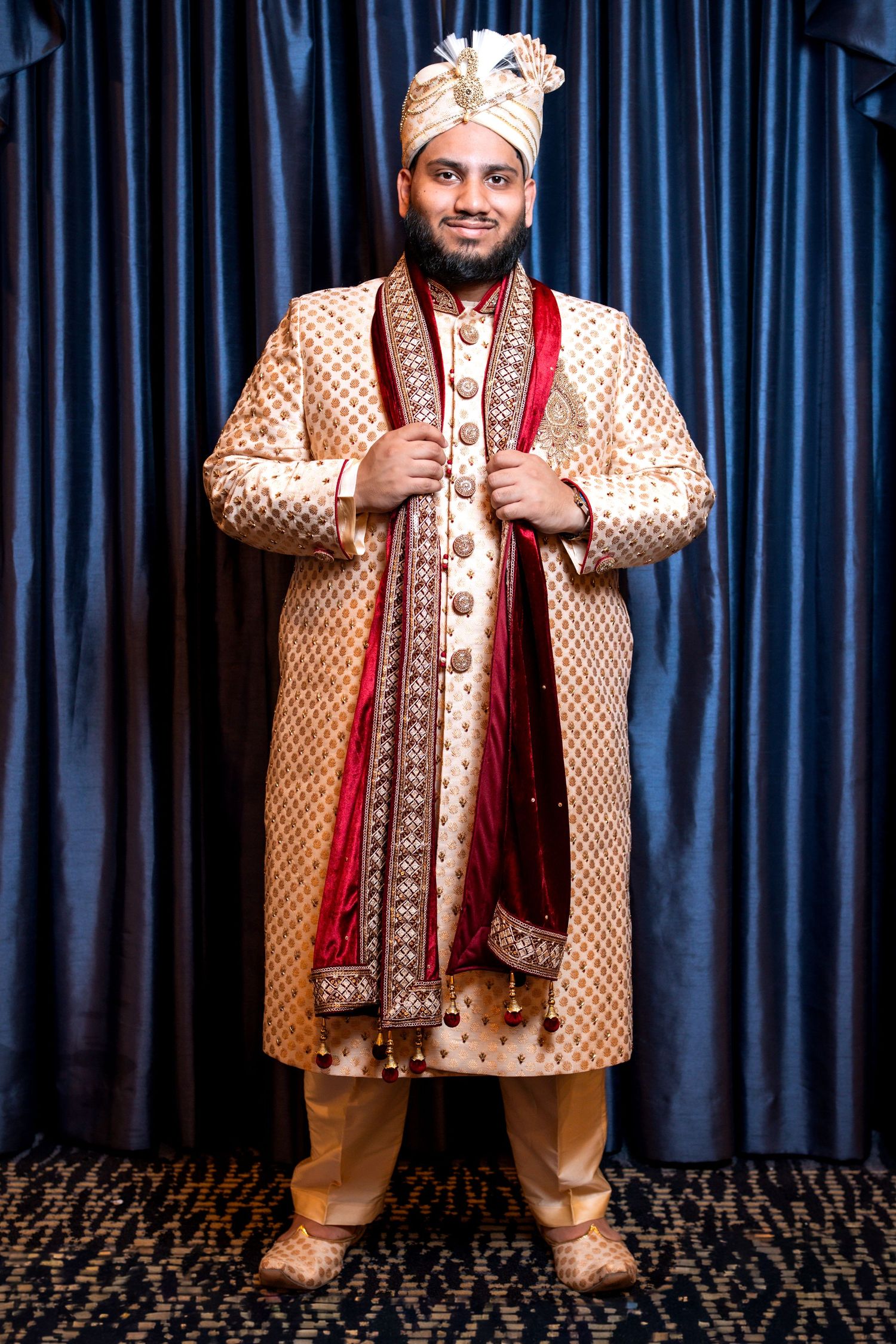 Indian groom in full traditional gold sherwani and turban standing proudly before blue curtains.