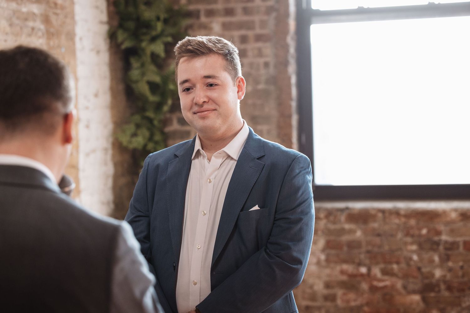 Young man in blue blazer and pink shirt smiling at someone in a rustic brick-walled event space.