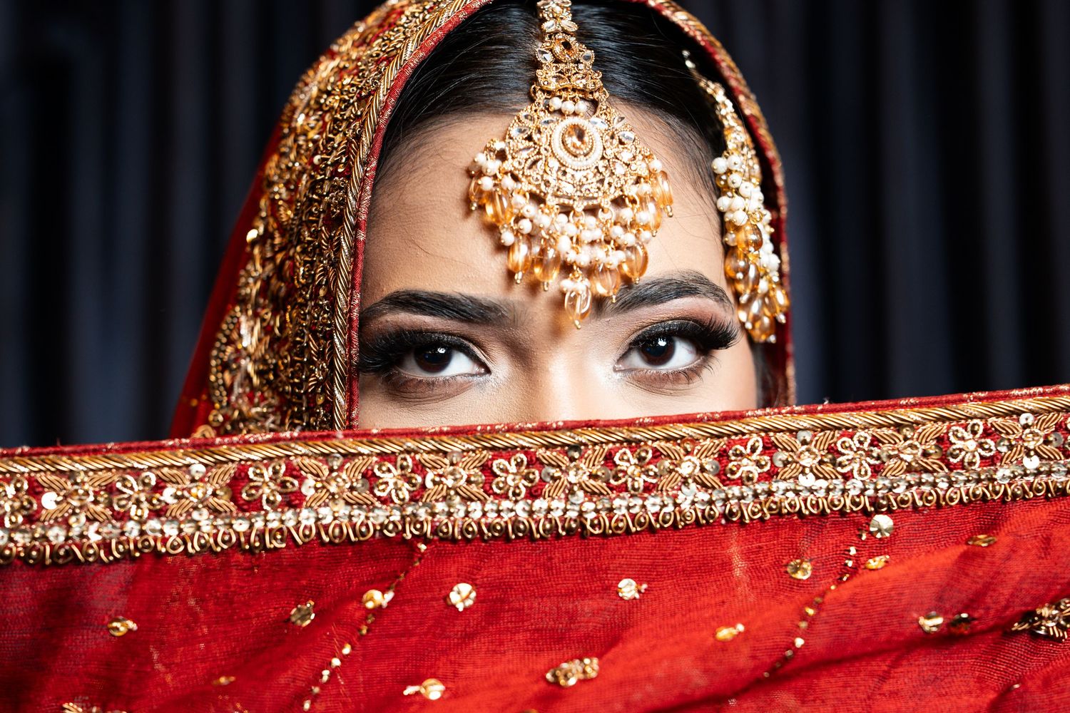 Indian bride in red and gold bridal attire with ornate maang tikka jewelry partially covering her face.