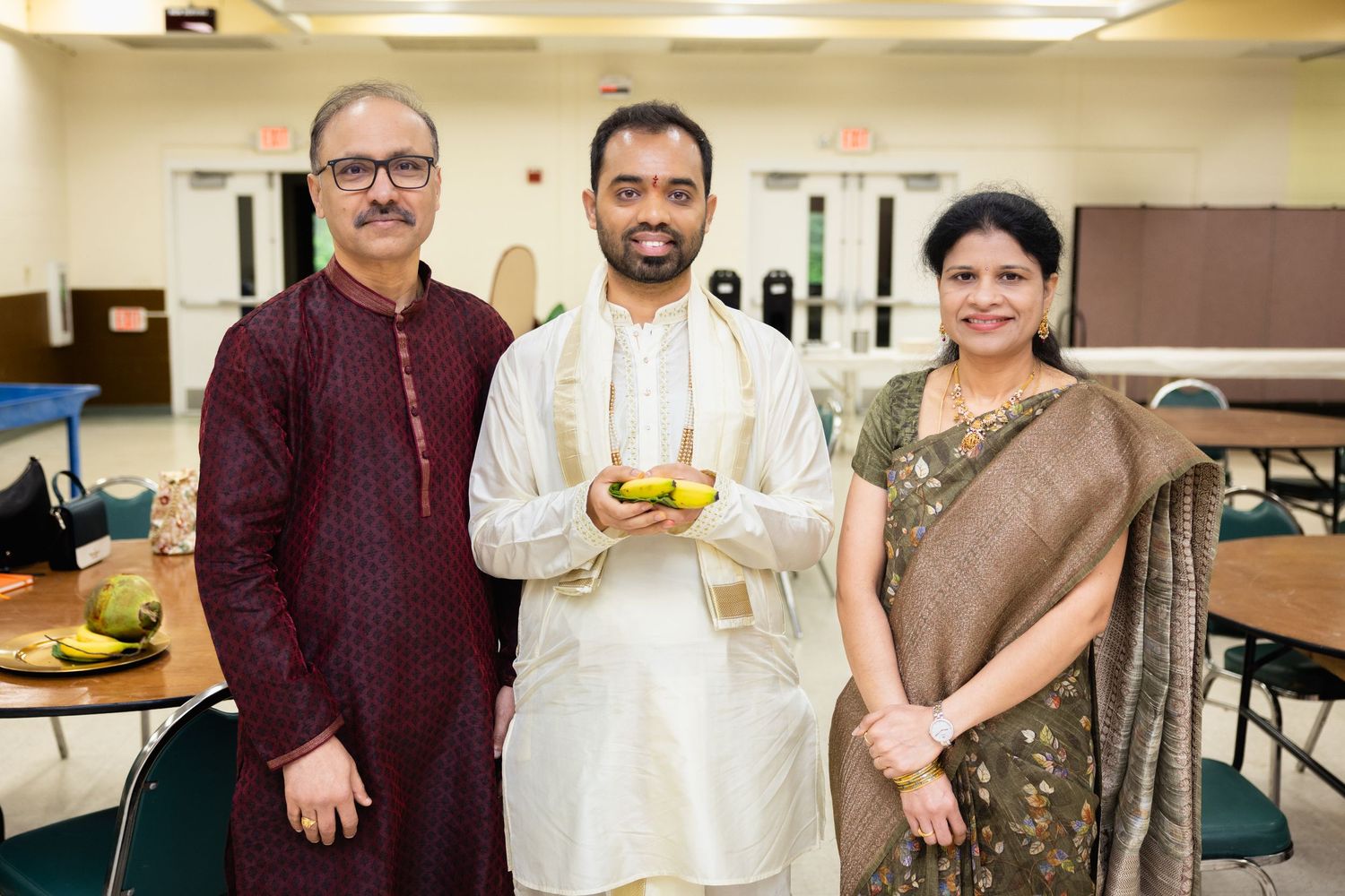 Indian groom in white kurta holding ceremonial bowl standing between parents in traditional attire indoors.