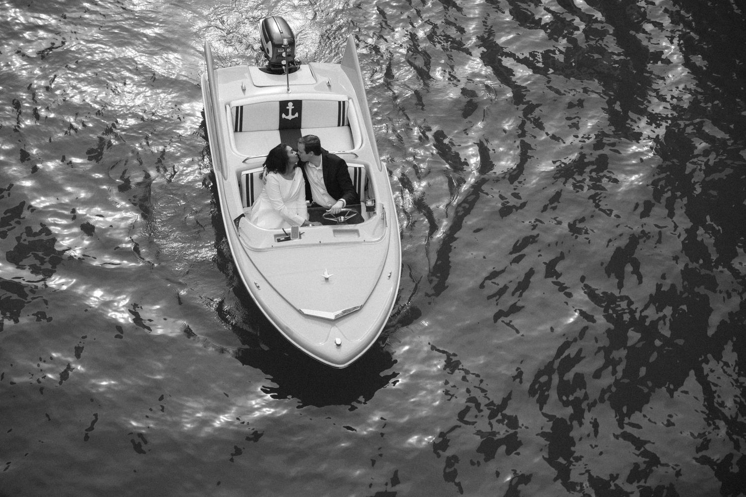 Aerial black and white shot of couple kissing in a white speedboat on shimmering water.