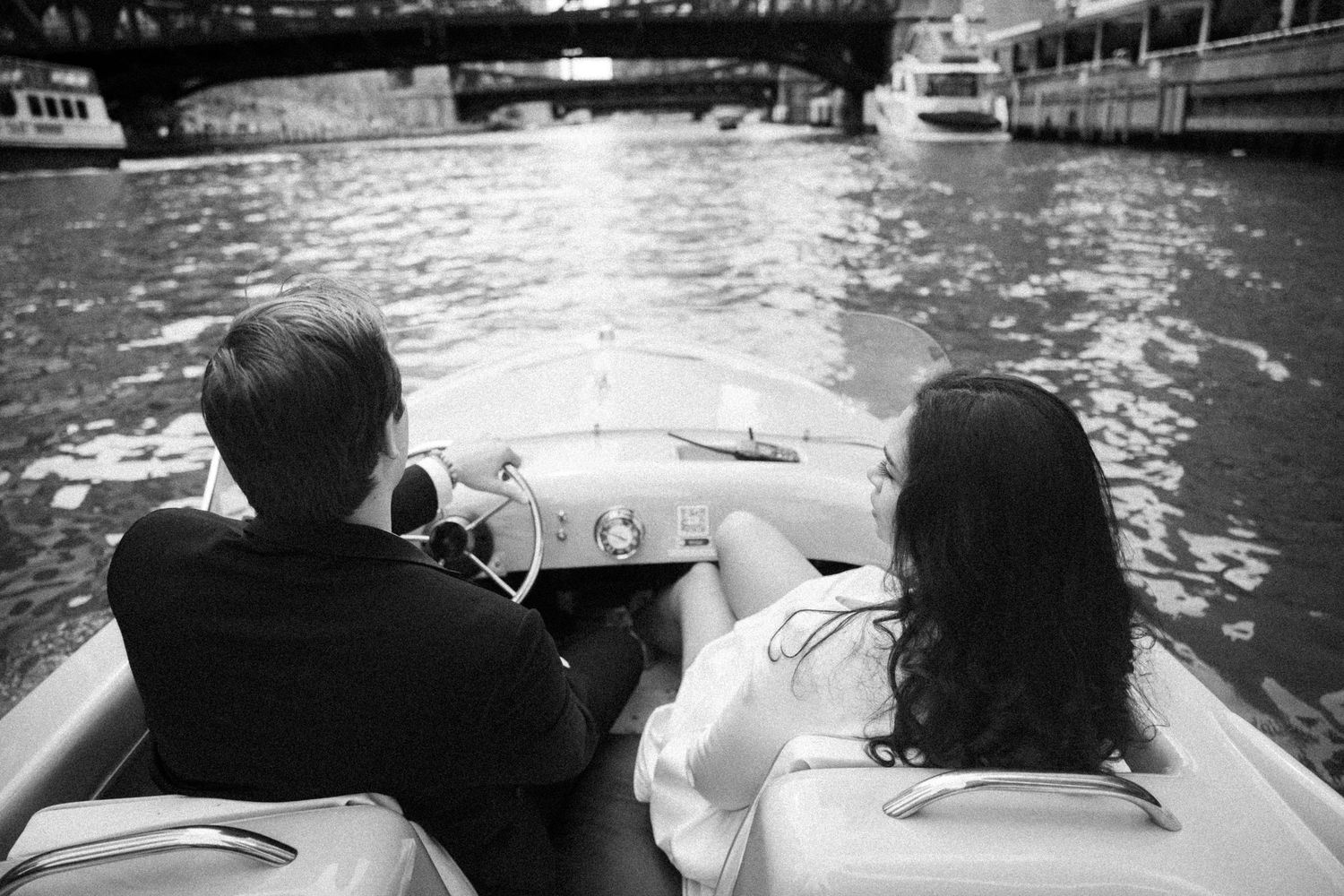 Black and white photo of a couple riding in a vintage speedboat on a city river with bridges in the background.