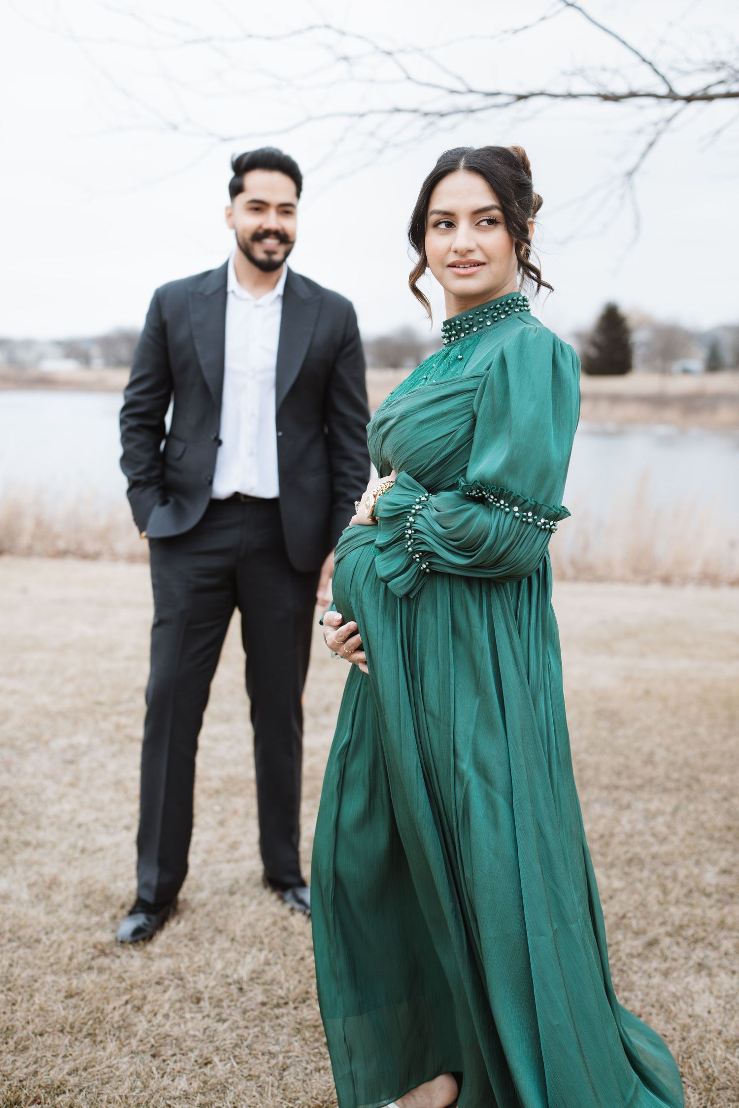 Expecting couple posing outdoors near lake, woman in stunning emerald green maternity gown.