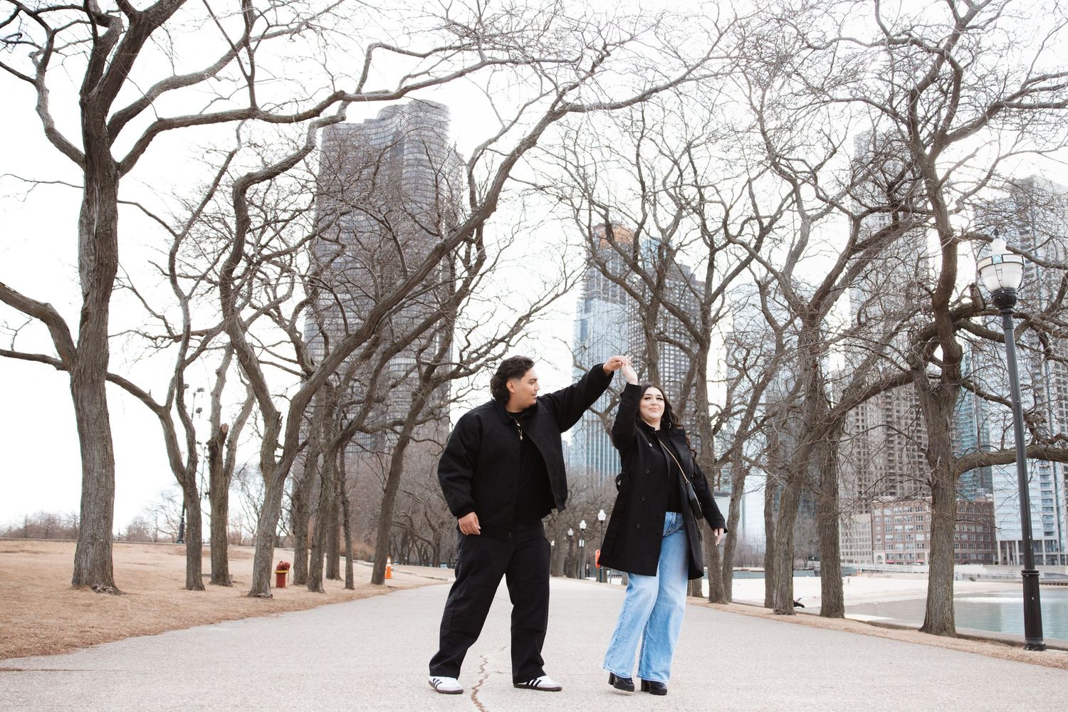 Couple dancing together on a tree-lined path in a winter park with city skyscrapers in the background.