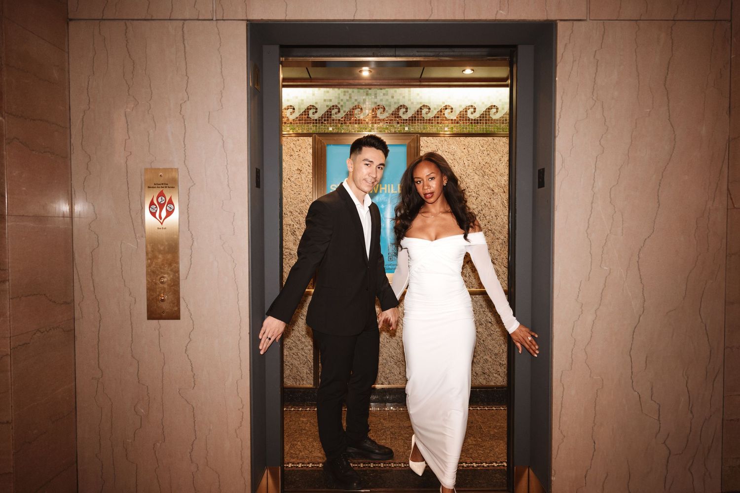 Stylish couple holding hands smiling in open elevator doorway, wearing black suit and white fitted gown.Chicago Wedding Photographer