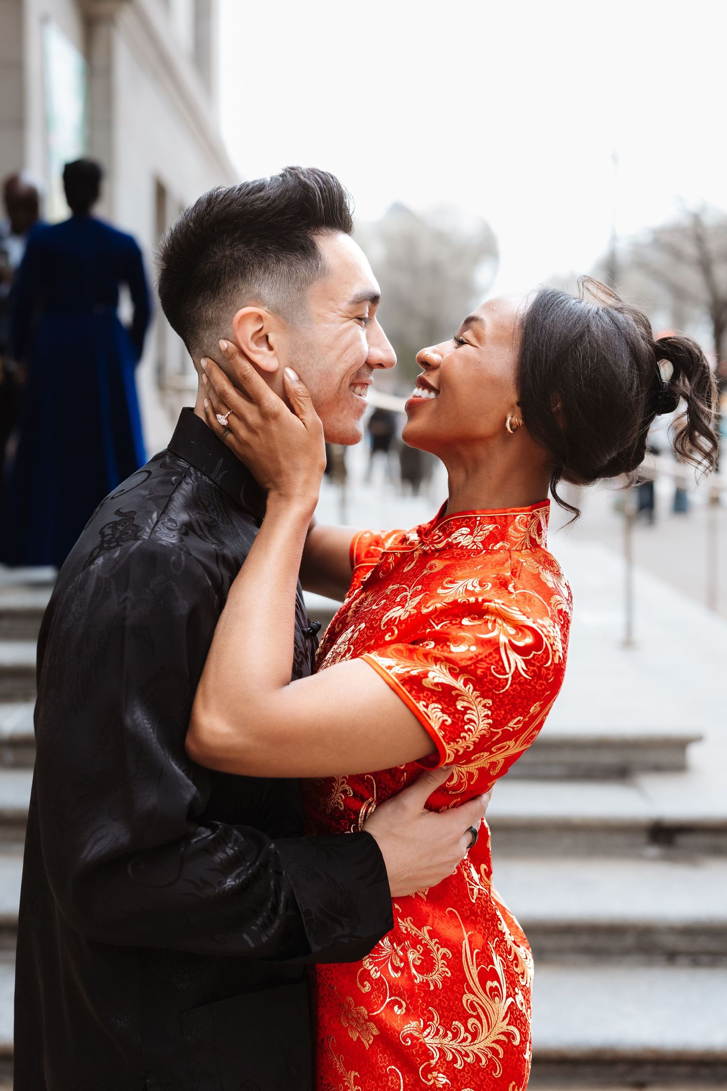 Couple in traditional Chinese attire embracing and smiling outdoors on steps, woman in red qipao, man in black.