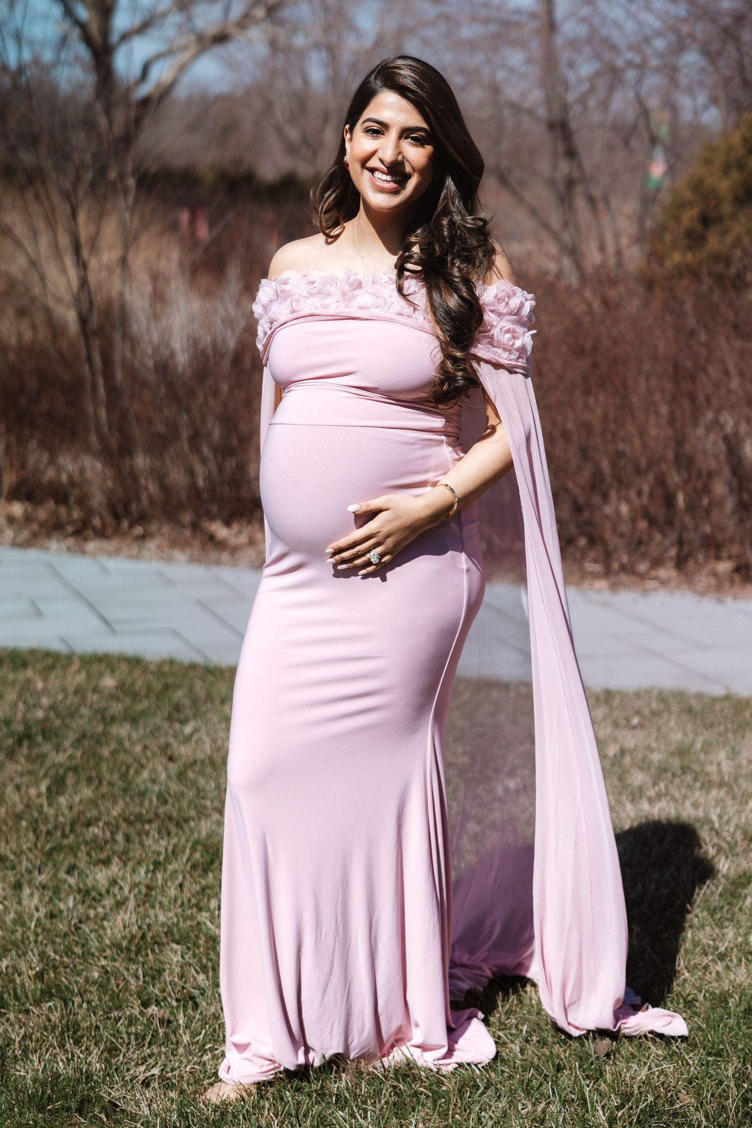 Smiling pregnant woman in elegant pink off-shoulder cape gown poses outdoors in spring garden.
