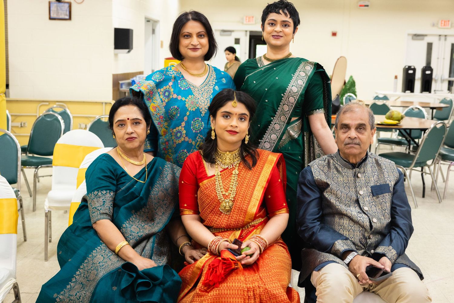 Indian family portrait with bride in red saree and gold jewelry surrounded by relatives in teal outfits.