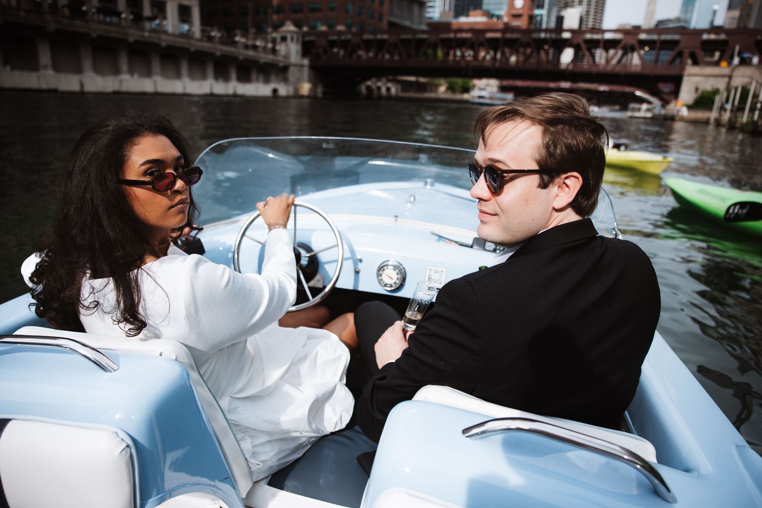 Stylish couple wearing sunglasses driving a classic blue speedboat along the Chicago River on a sunny day.