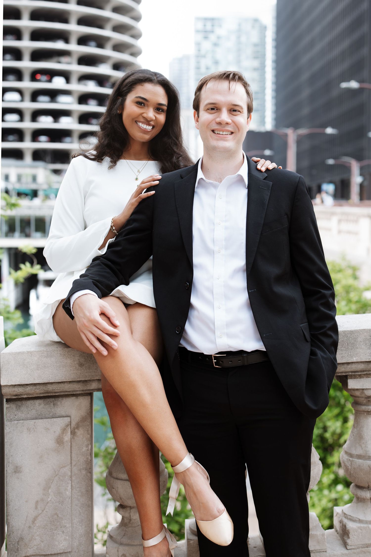Interracial couple in formal attire smiling together on urban bridge with city skyline background.