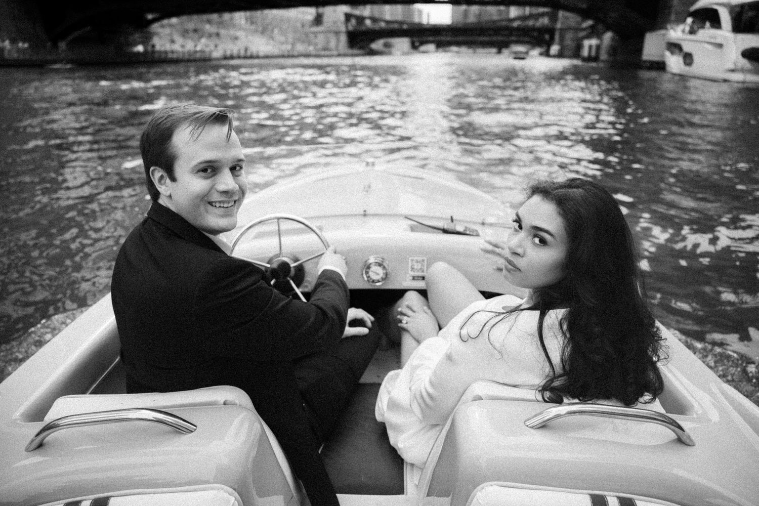 Black and white photo of a couple sitting in a vintage speedboat on a river, smiling at the camera.