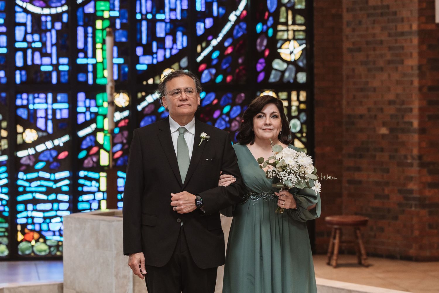 Elegant couple posing in church with stained glass windows, woman holding bouquet in teal dress beside suited man.