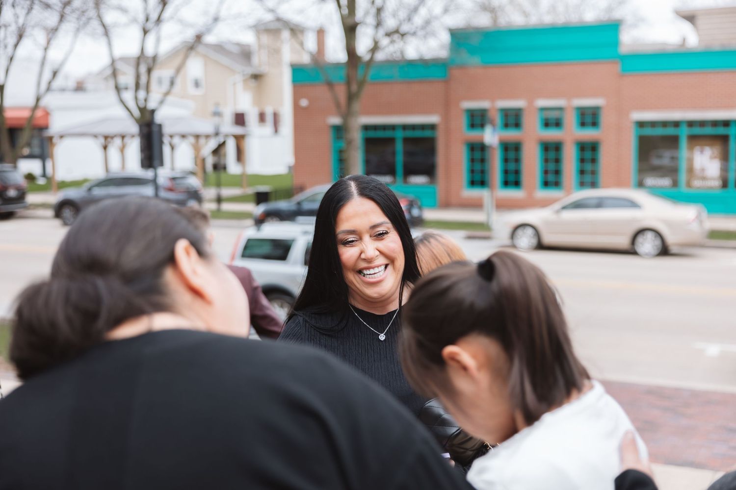 A smiling young woman with long black hair laughs with friends outdoors near a teal storefront.