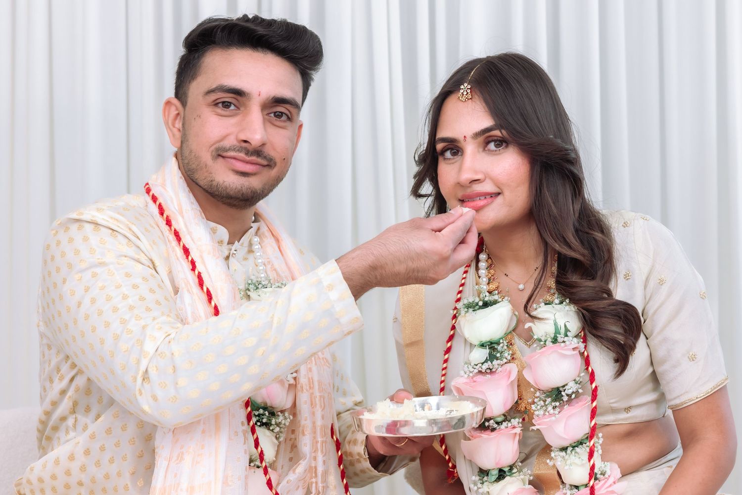 Indian groom in white sherwani feeds bride during traditional wedding ceremony while both wear colorful floral garlands.