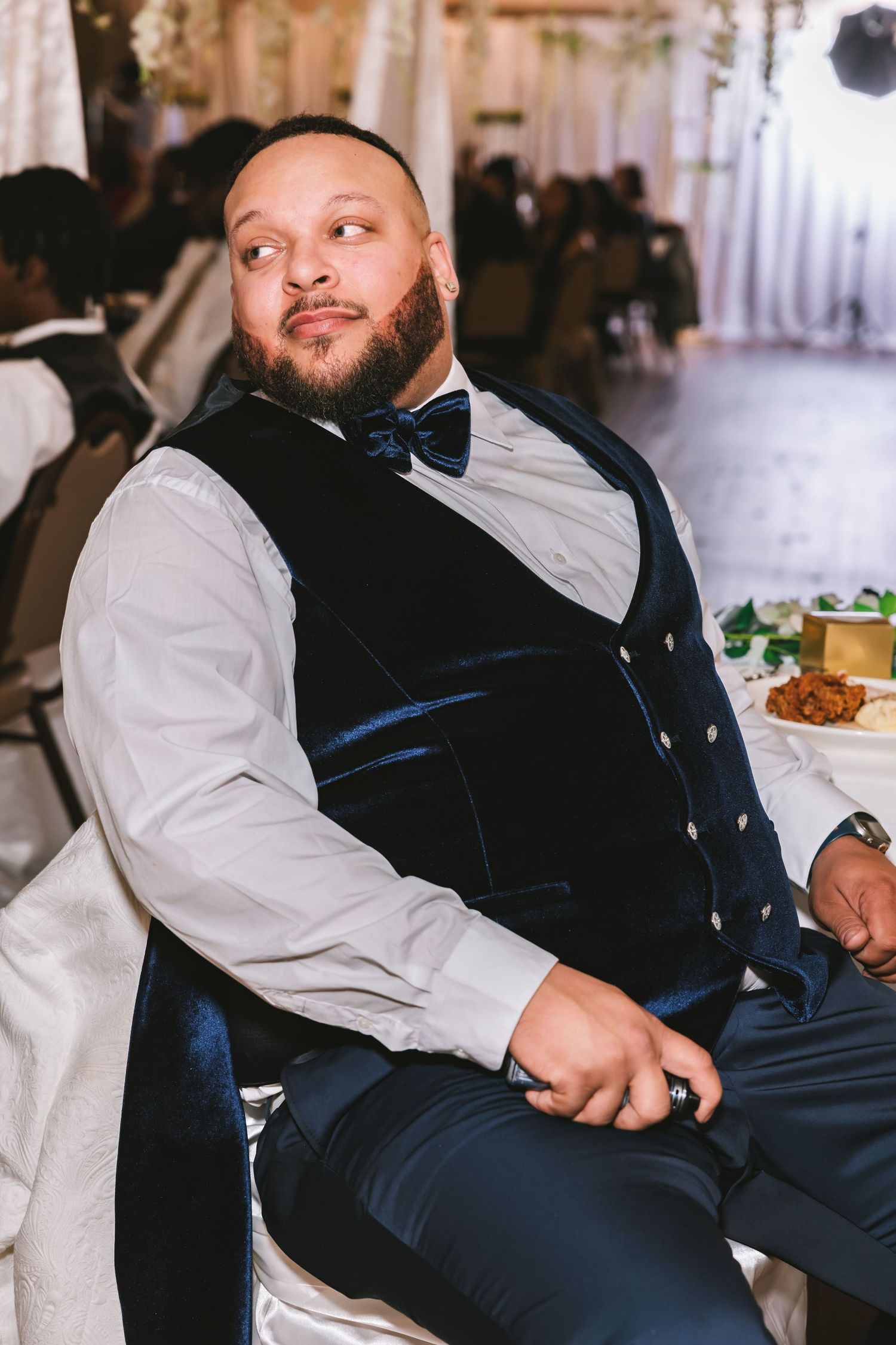 Groom in navy velvet vest and bow tie sits relaxed at wedding reception dinner table.