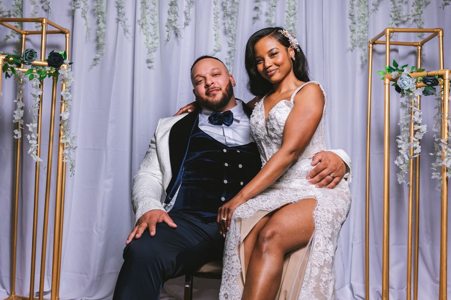 Bride and groom posing at white draped backdrop with gold floral stands at wedding reception.