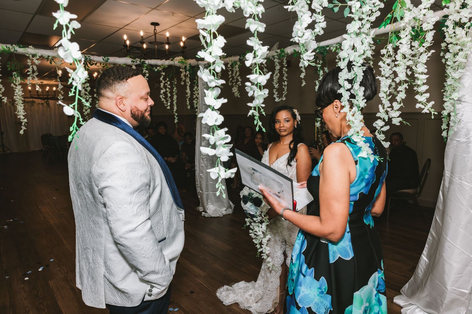 Wedding officiant reads vows to bride and groom standing beneath elegant hanging white floral greenery arch.