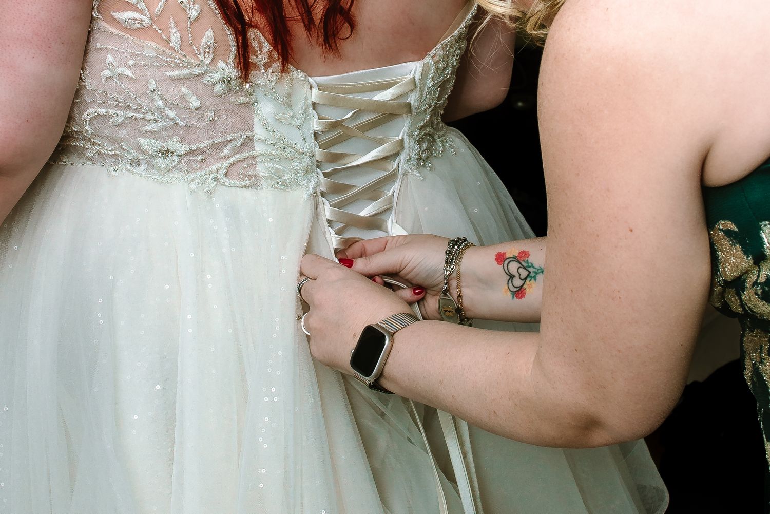 Close-up of wedding dress corset lacing being tied by bridesmaid wearing an Apple Watch and floral bracelet.