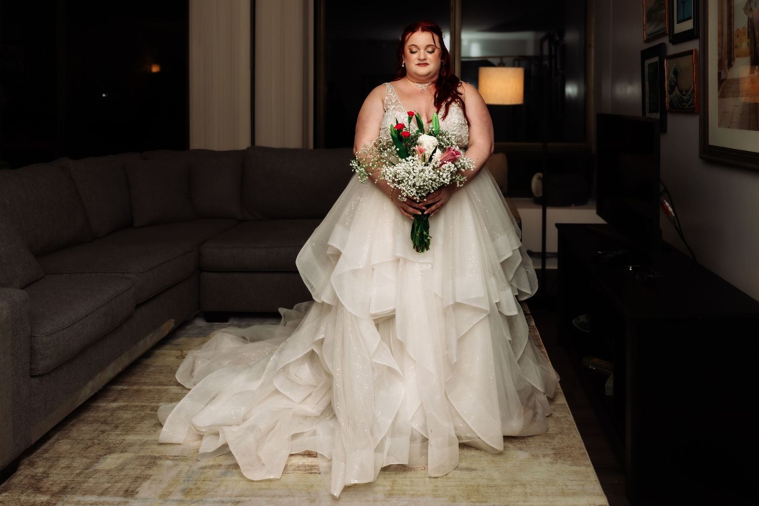Full-length portrait of bride in ruffled tulle ball gown holding floral bouquet in dimly lit room.