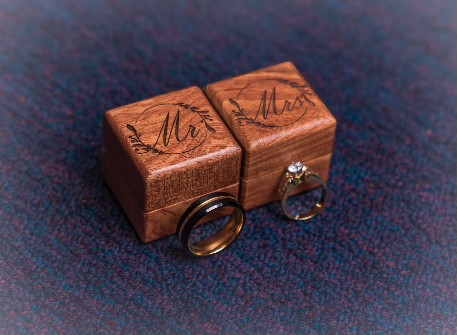 Two wooden Mr. and Mrs. ring boxes with engraved calligraphy displayed alongside wedding bands on a textured surface.