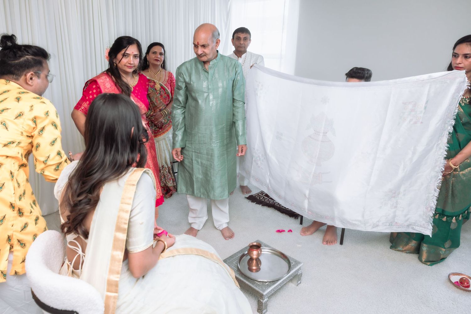 Family holds white cloth during traditional Indian wedding ritual with bride seated in white lehenga.