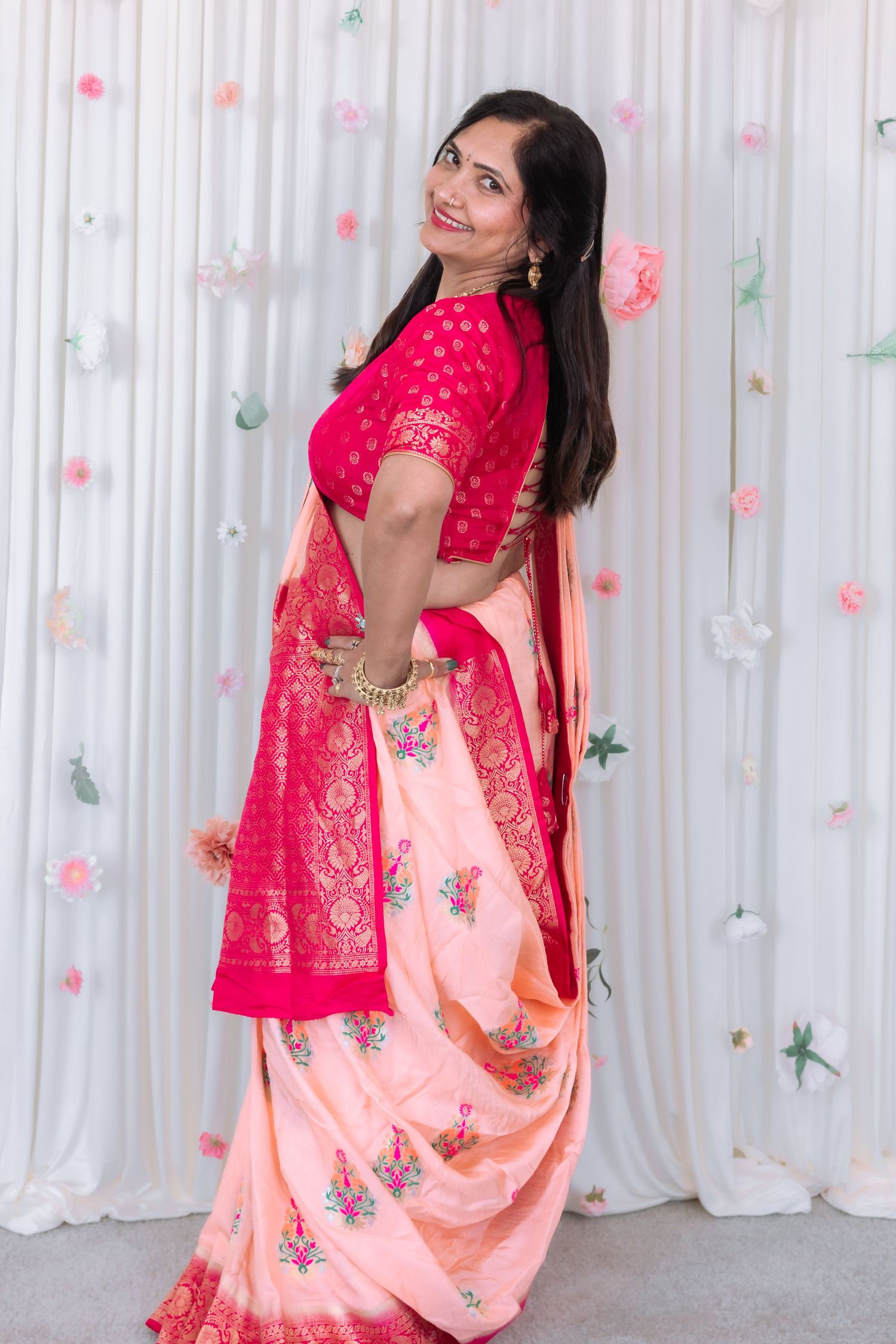 Indian woman in vibrant pink red Banarasi saree poses gracefully at floral wedding ceremony backdrop.