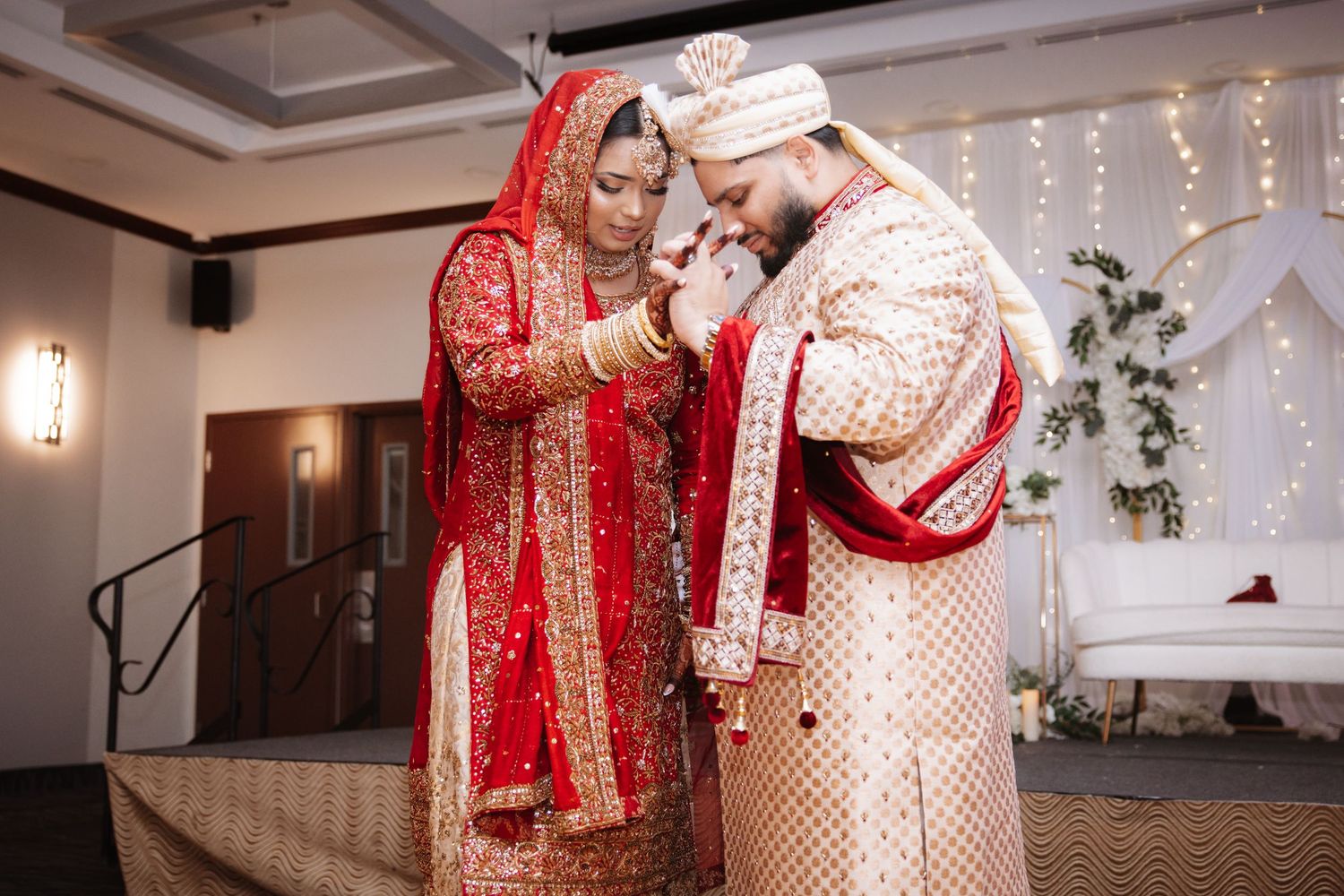 Indian bride in red lehenga and groom in gold sherwani exchanging garlands at wedding ceremony.