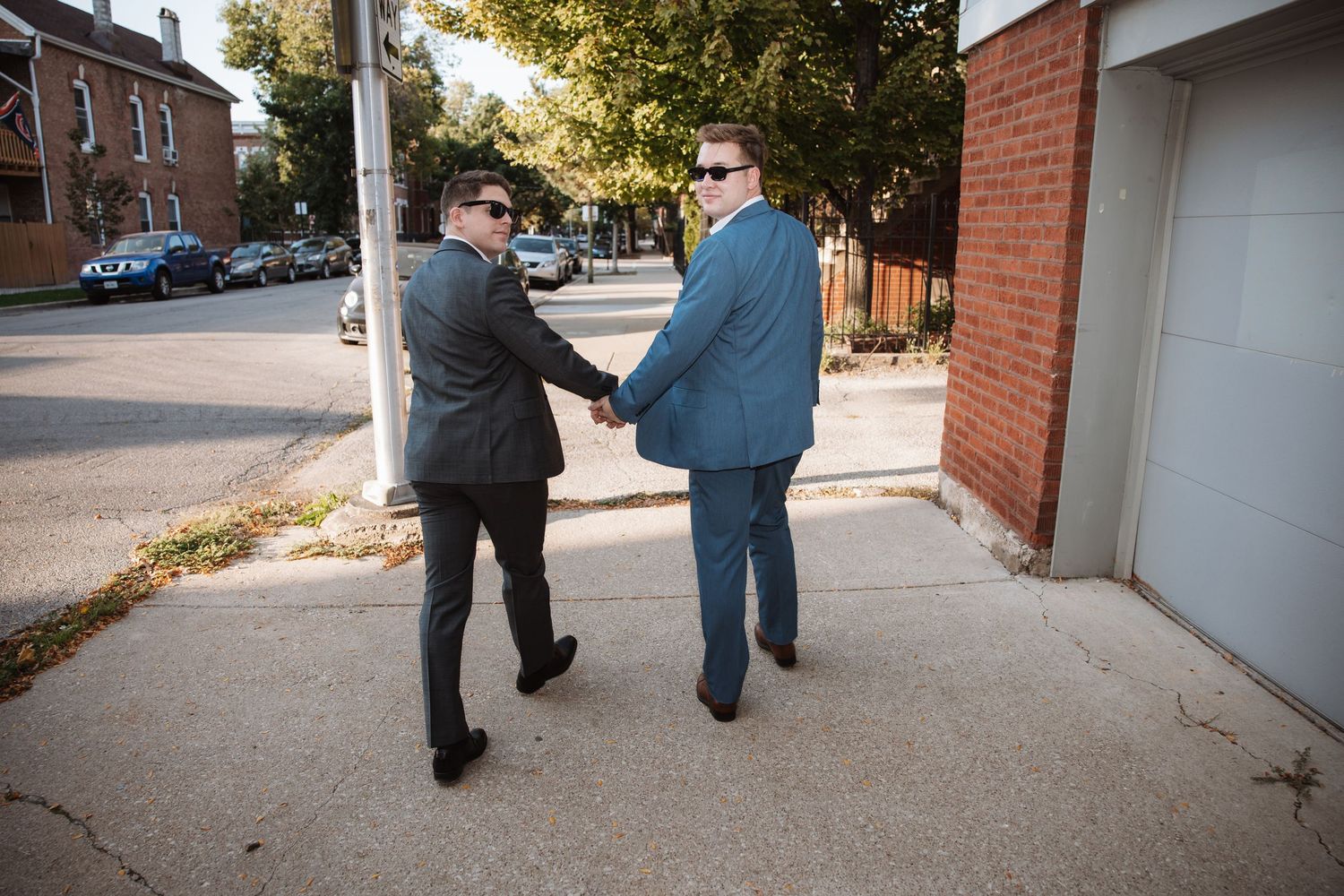 Two grooms in dark and blue suits shake hands while smiling on a sunny city street.