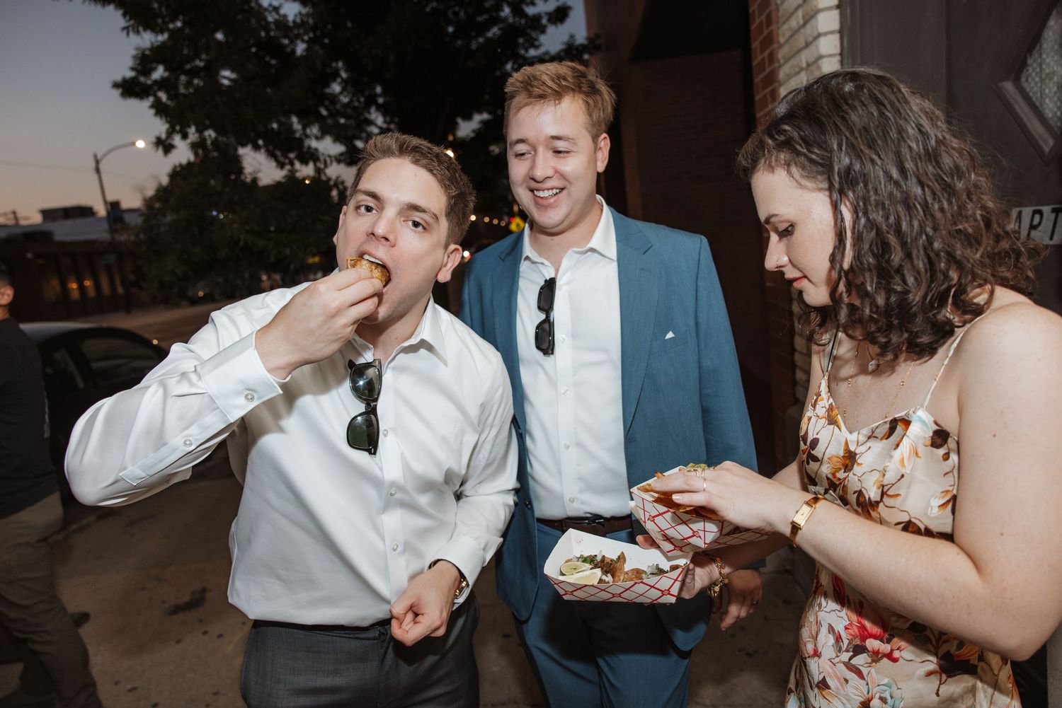 Three guests enjoying food outdoors at a wedding reception at dusk.