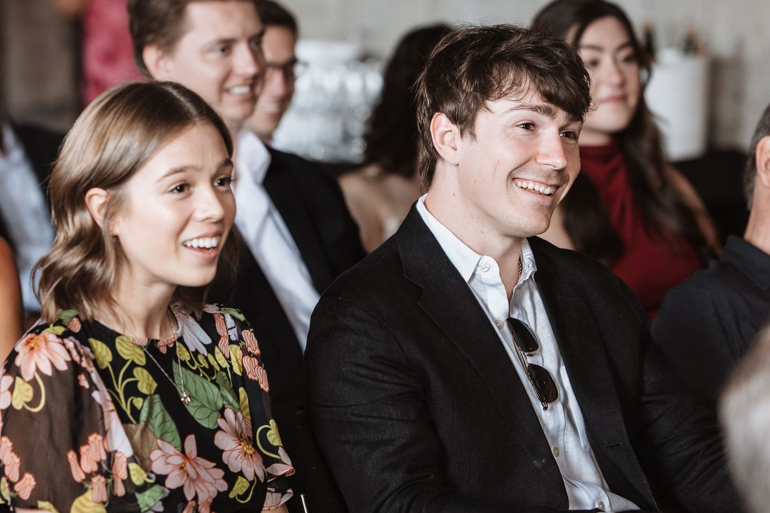 Smiling young couple seated among guests at a wedding ceremony in a bright venue.