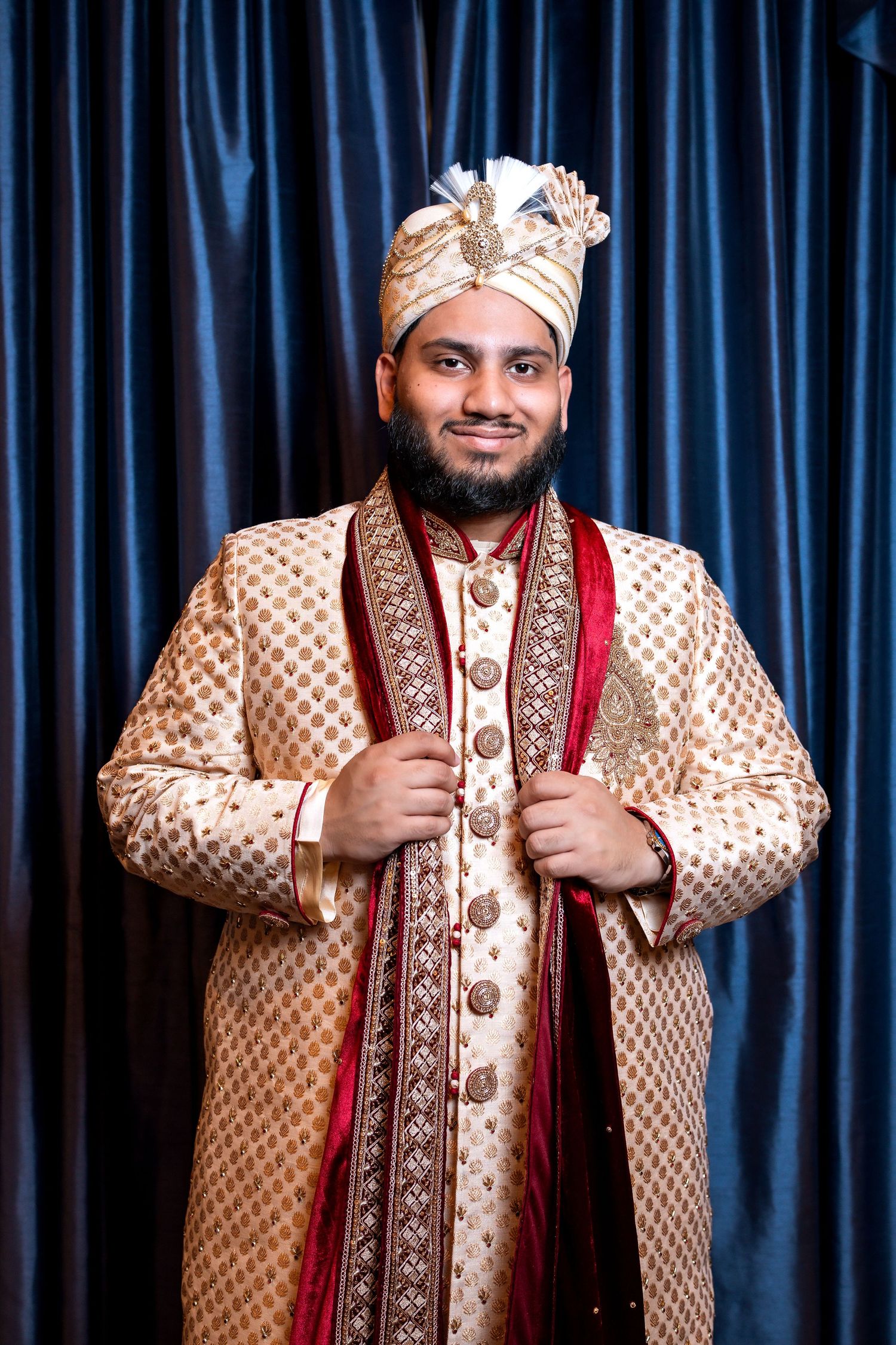 Smiling Indian groom in embroidered gold sherwani and white turban posing before blue curtains.