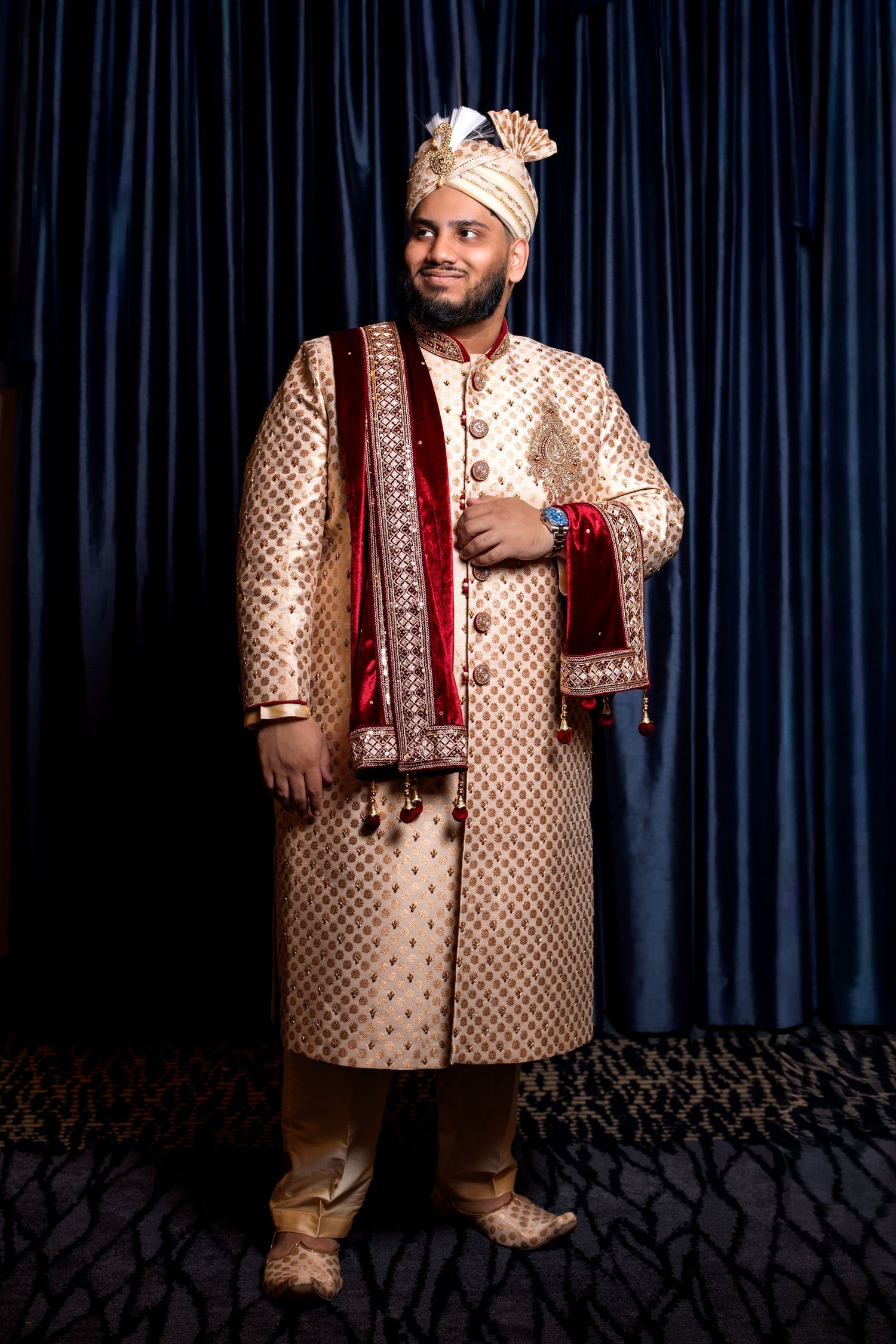 South Asian groom in traditional gold sherwani and ornate turban posing against rich blue velvet curtains.