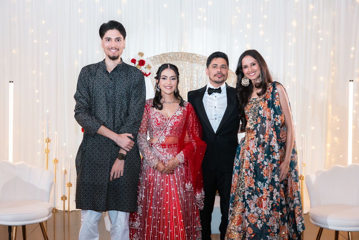 Four guests posing with bride in red lehenga and groom in tuxedo at Indian wedding reception.