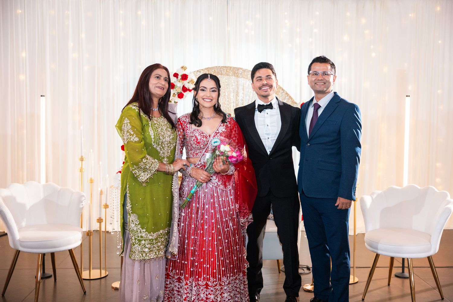 Bride in red lehenga stands with groom in tuxedo and two guests at decorated wedding venue.