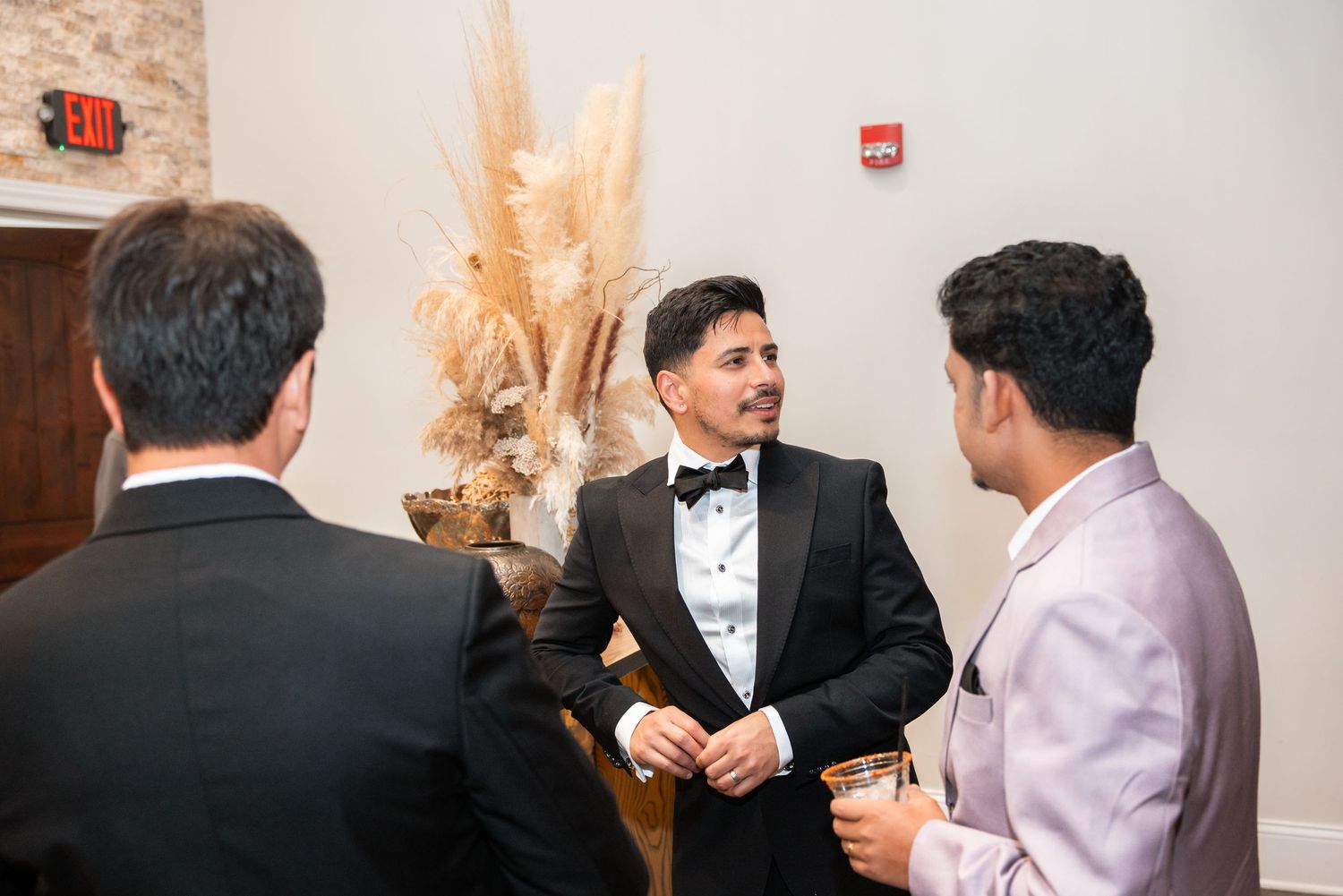 Man in tuxedo socializes with guests at an elegant formal event near pampas grass decor.