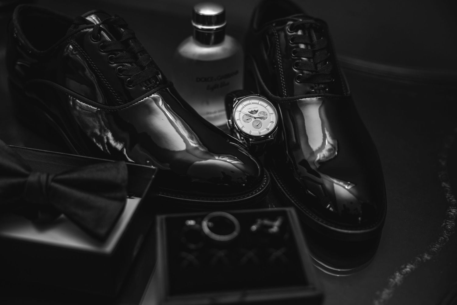 Black and white flat lay of groom's patent shoes, watch, and bow tie.