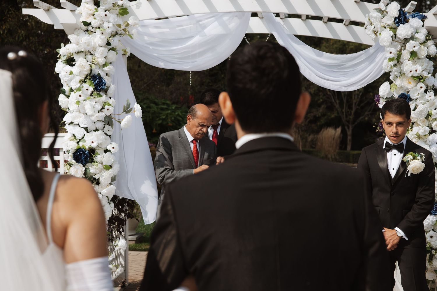 Wedding ceremony at a floral arch with officiant, groom, and guests gathered outdoors.