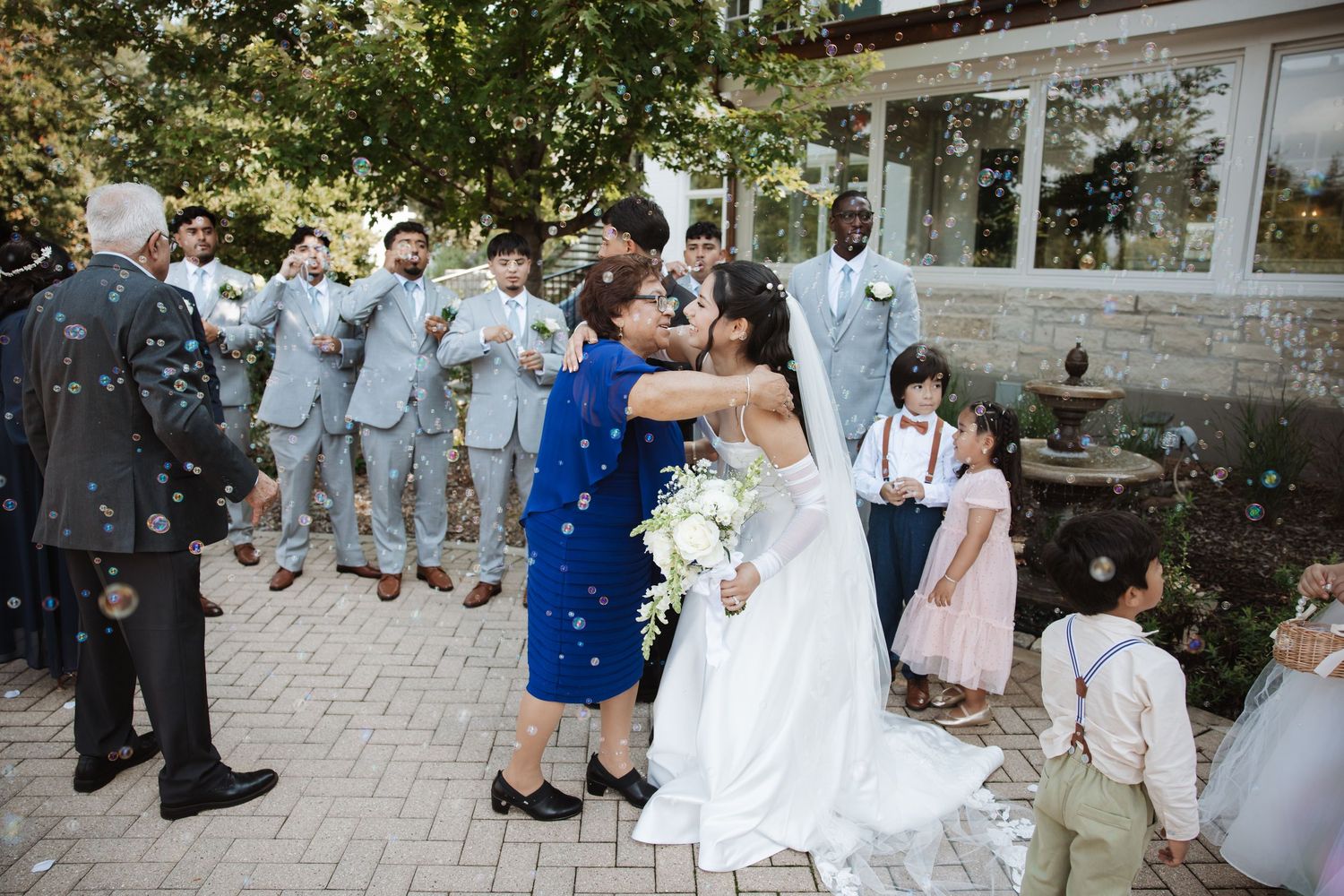 Bride and groom kissing while a woman in blue embraces them surrounded by wedding party and children.