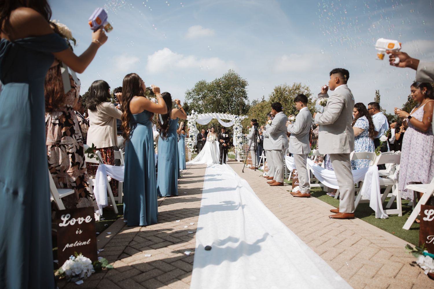 Outdoor wedding ceremony aisle with bridesmaids in blue dresses and groomsmen in gray suits lining the path.