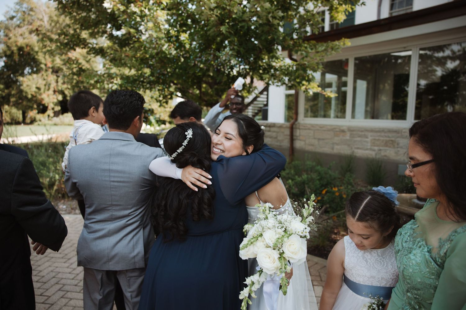 Bride hugging a female guest in blue dress while holding white bouquet at outdoor wedding reception.