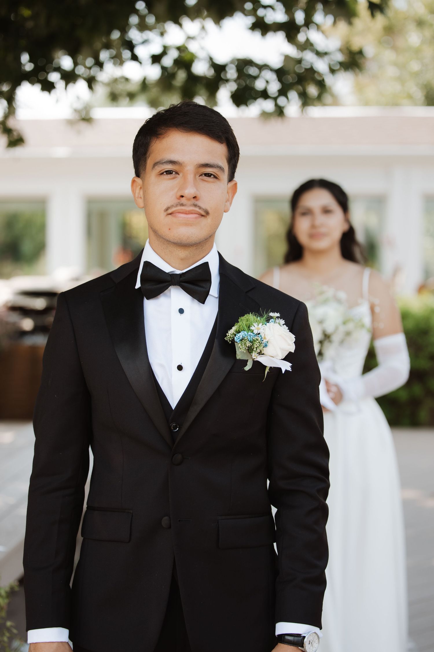 Groom in black tuxedo with boutonniere stands as bride in white gown poses behind him.
