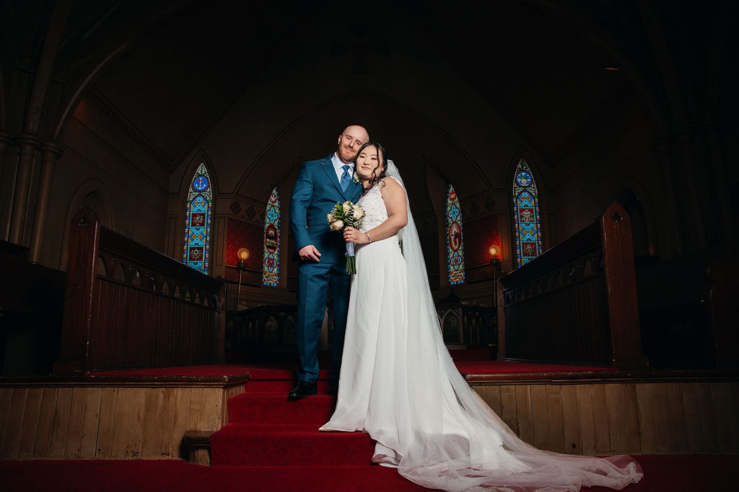 Bride and groom pose together at the altar of a wooden church with red carpet and stained glass windows.