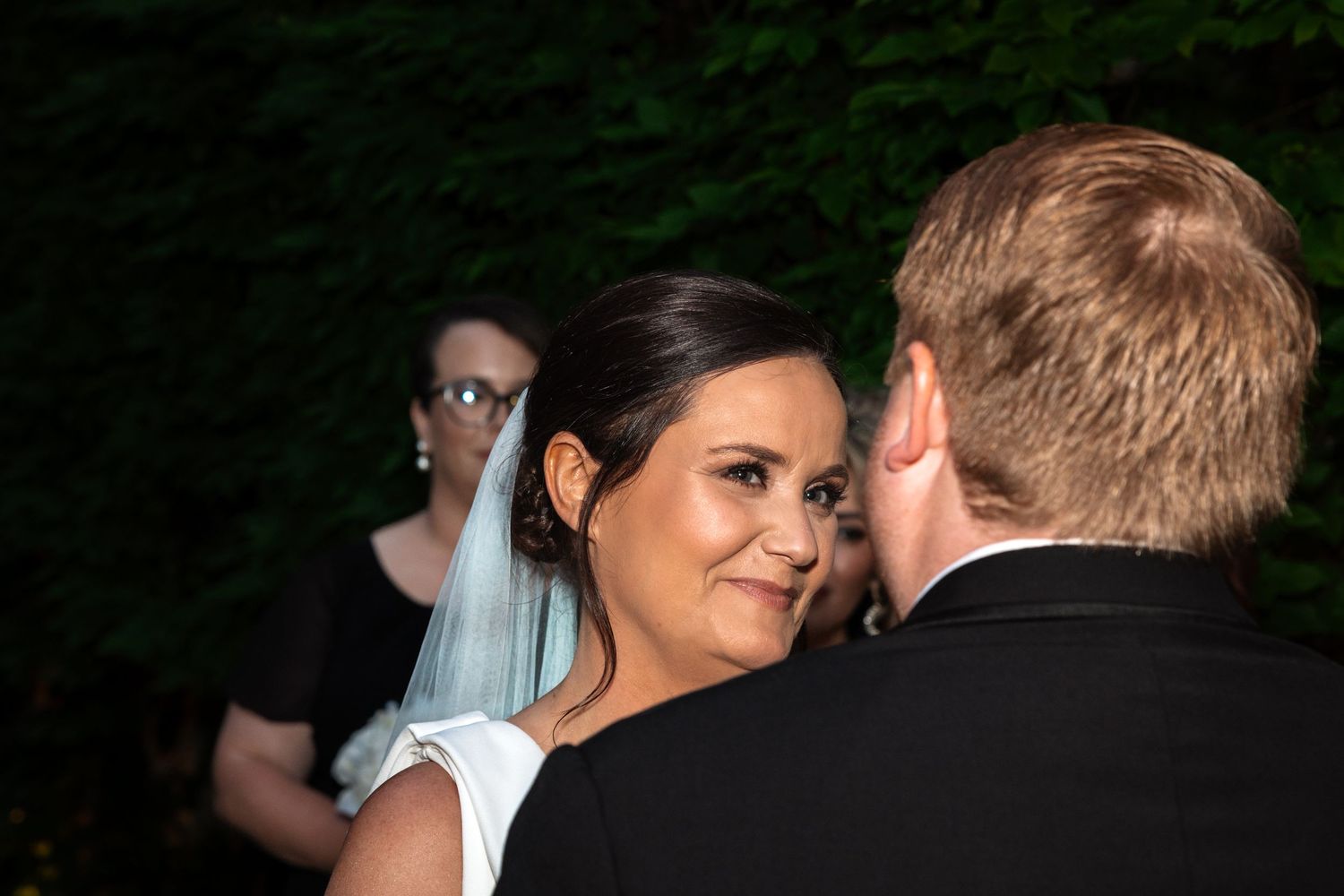 Smiling bride in white dress and blue veil gazes at groom during outdoor wedding ceremony at night.