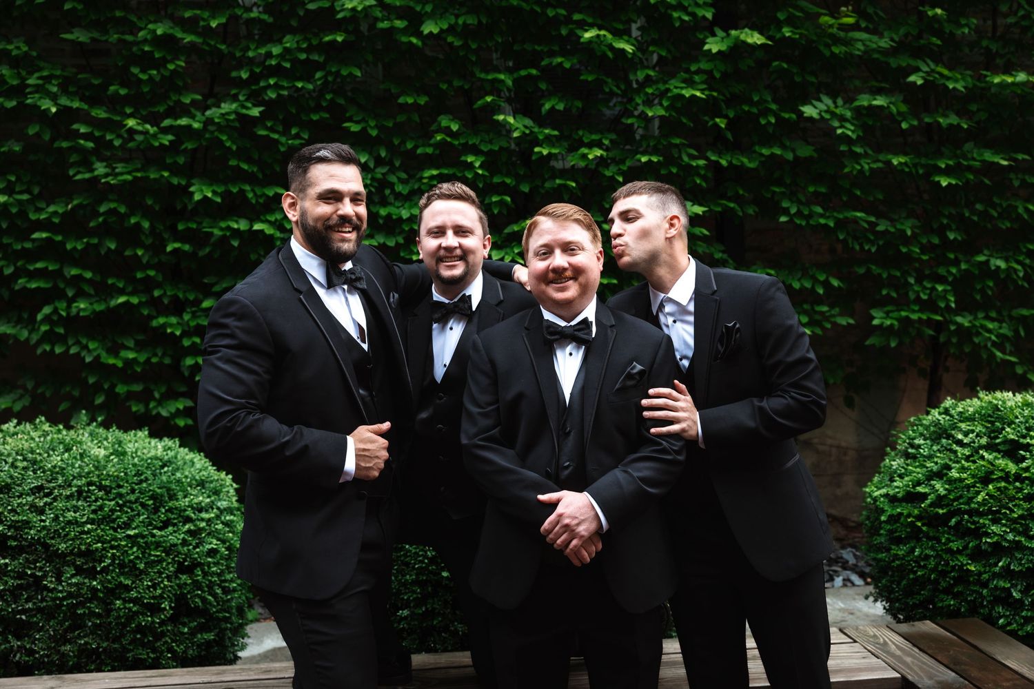 Four groomsmen in black tuxedos laughing and joking together outdoors against a lush green ivy backdrop.