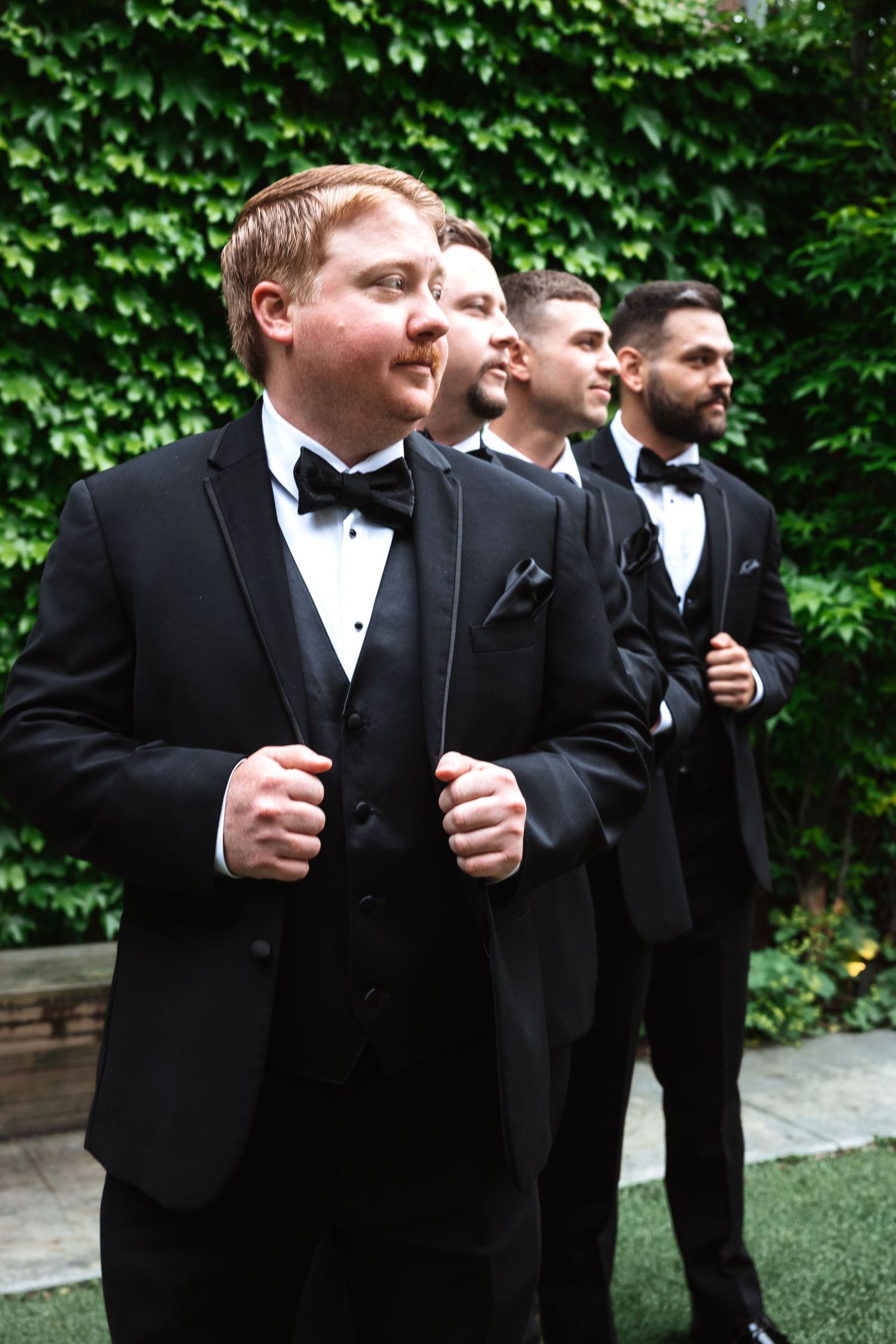 Groom and groomsmen in black tuxedos standing in a diagonal line outdoors against a green ivy-covered wall.