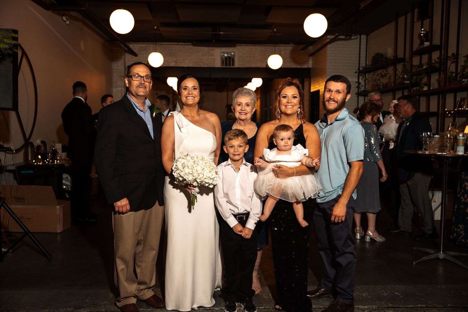 Bride in white gown poses with family group including children at wedding reception venue.